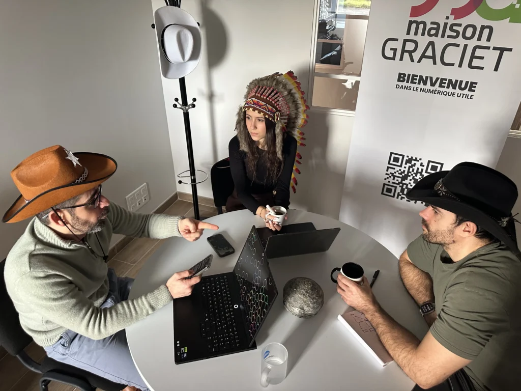 Manon Delcambre, Stéphane Graciet et Valentin Charrier portant des chapeaux de cow-boy et une coiffe à plumes en réunion de travail autour d'une table à côté d'un kakémono "maison GRACIET - Bienvenue dans le numérique utile". Stéphane au chapeau de shérif à gauche pointe l'écran de son ordinateur.