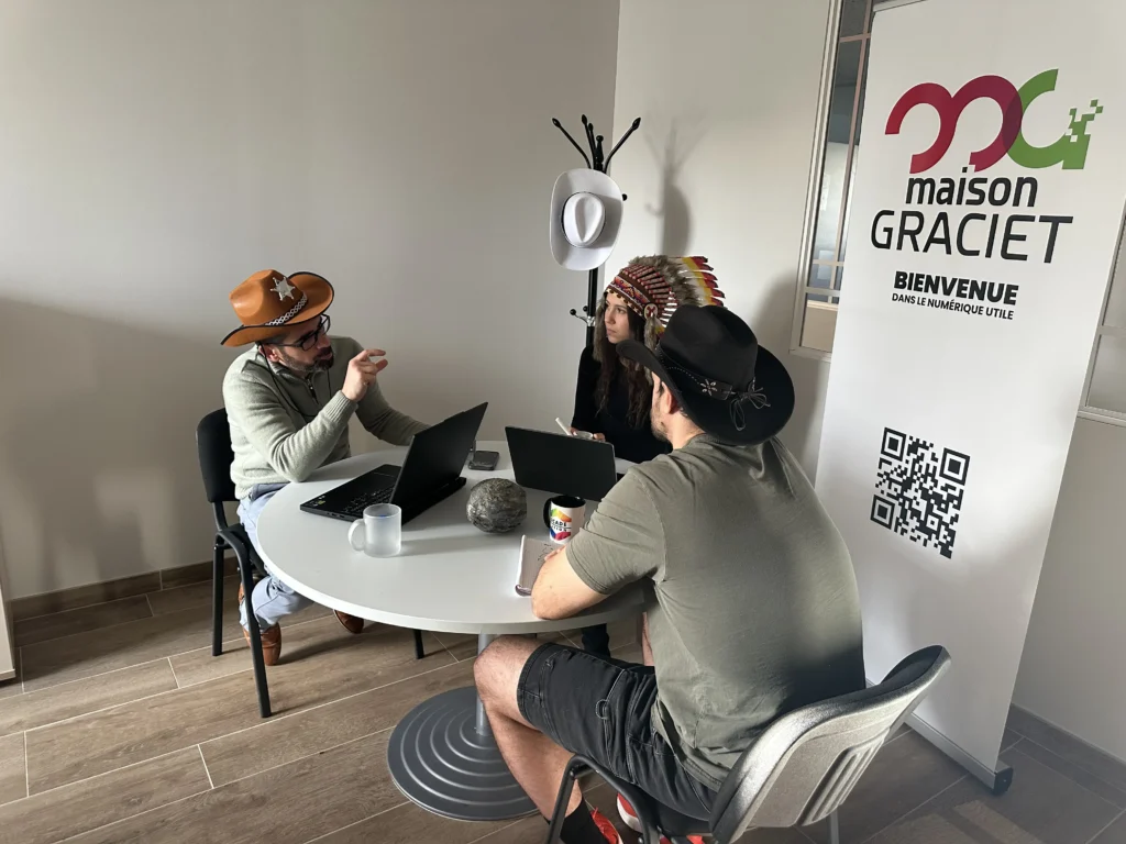 Manon Delcambre, Stéphane Graciet et Valentin Charrier portant des chapeaux de cow-boy et une coiffe à plumes en réunion de travail autour d'une table à côté d'un kakémono "maison GRACIET - Bienvenue dans le numérique utile". Stéphane au chapeau de shérif argumente avec les mains pour expliquer un concept à ses deux collègues attentifs.